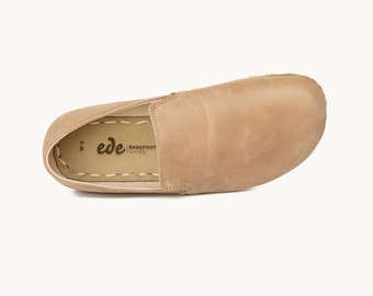 May include: Tan slip-on shoe with a rounded toe. The shoe is made of a soft, textured material. The brand name "ede BAREFOOT SHOES" is printed on the insole. The shoe is a size 9W.