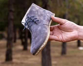 May include: A gray suede boot with blue laces is held up against a blurred forest background. The boot has a stitched sole and a light-colored interior. The leather has a textured appearance, and the laces are a darker shade of blue.
