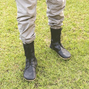 May include: A person wearing black leather boots with laces, standing on green grass. The boots have a rounded toe and a textured sole. The person is also wearing light gray pants.