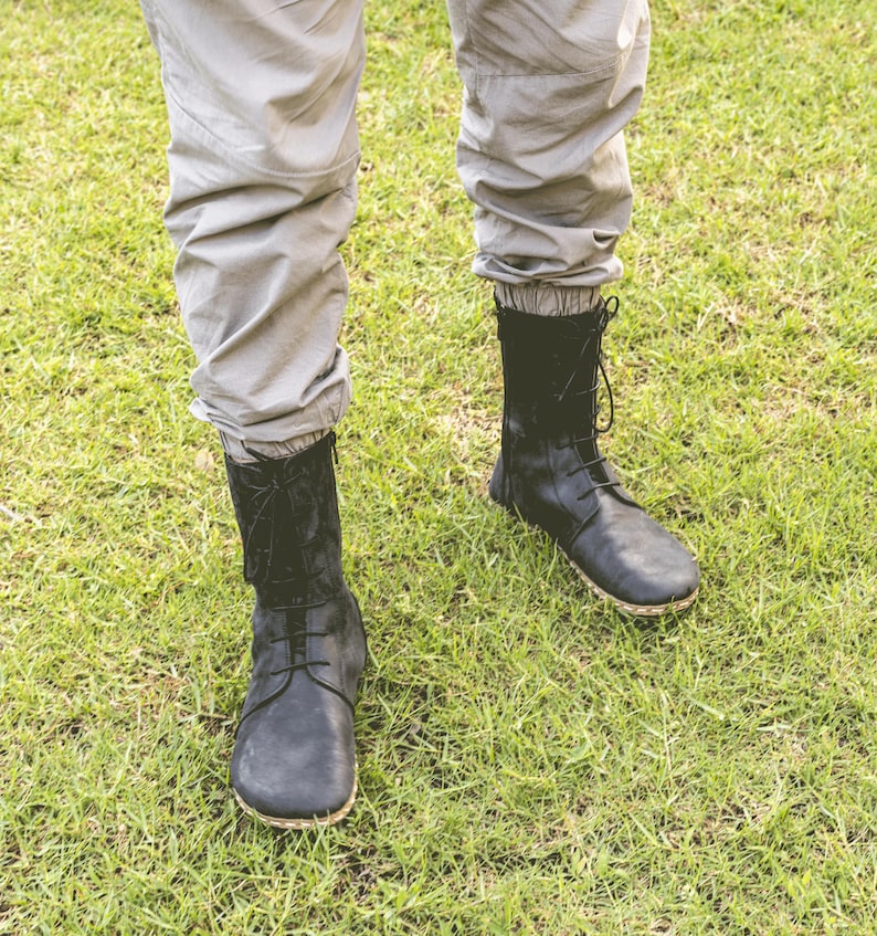 May include: A person wearing black leather boots with laces, standing on green grass. The boots have a rounded toe and a textured sole. The person is also wearing light gray pants.