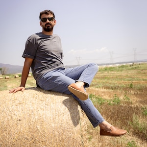 May include: A person wearing a gray t-shirt, blue jeans, and brown leather loafers sits on a large bale of hay in a field. The person is wearing sunglasses and the background shows a clear blue sky and power lines.