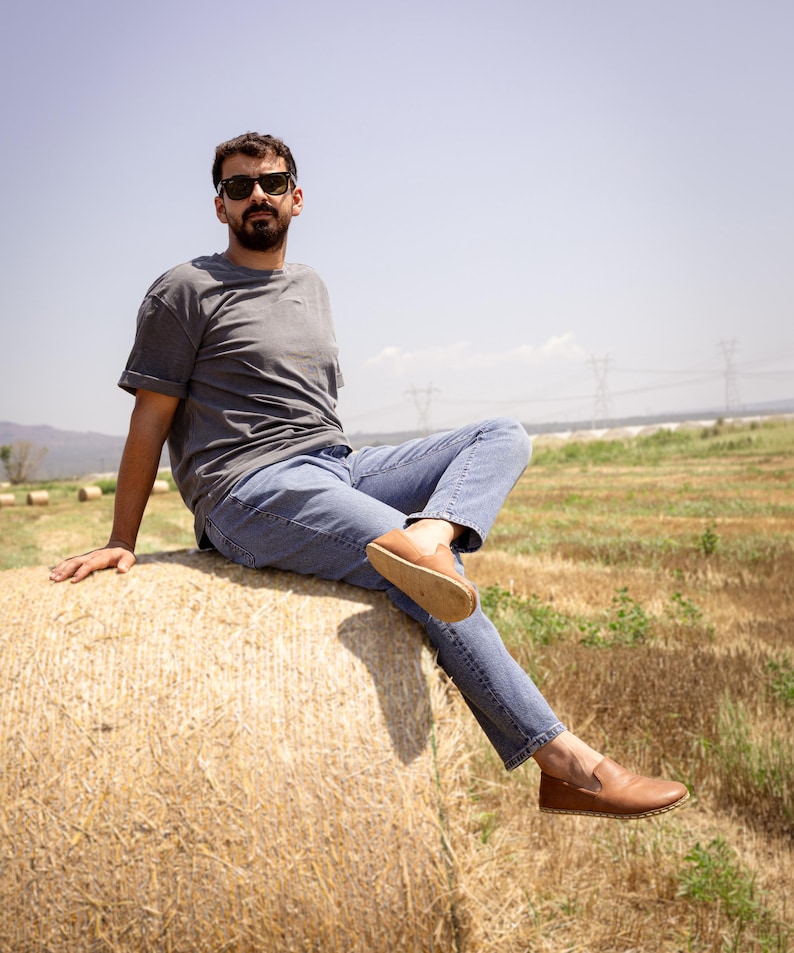 May include: A person wearing a gray t-shirt, blue jeans, and brown leather loafers sits on a large bale of hay in a field. The person is wearing sunglasses and the background shows a clear blue sky and power lines.