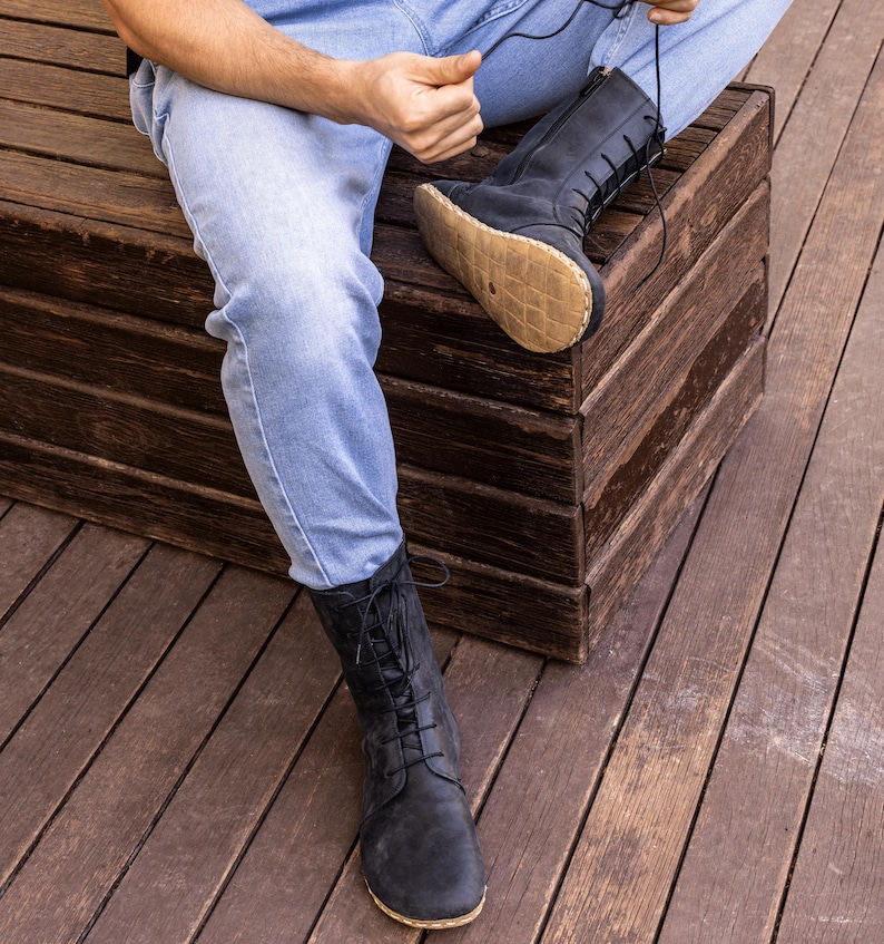 May include: Black leather lace-up boots with a side zipper. The boots have a natural-colored sole and are worn with blue jeans. The boots are mid-calf height and have a classic, rugged design.