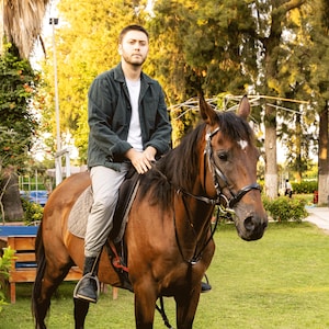 May include: A person sitting on a brown horse with a black bridle and saddle. The horse is in a grassy area with trees in the background. The person is wearing a dark green jacket, white shirt, and light gray pants.