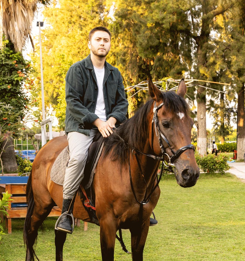 May include: A person sitting on a brown horse with a black bridle and saddle. The horse is in a grassy area with trees in the background. The person is wearing a dark green jacket, white shirt, and light gray pants.