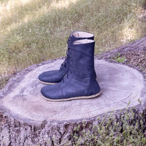 May include: A pair of navy blue leather boots with a lace-up design. The boots have a mid-calf height and a stitched sole. They are displayed on a tree stump outdoors.