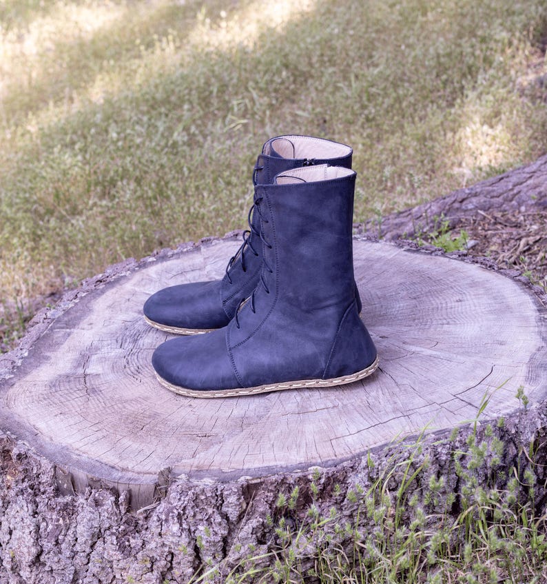 May include: A pair of navy blue leather boots with a lace-up design. The boots have a mid-calf height and a stitched sole. They are displayed on a tree stump outdoors.