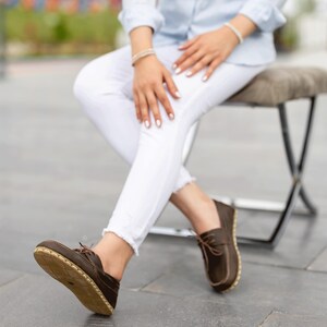 May include: Brown leather boat shoes with light brown stitching and laces. The soles are a light tan color. The shoes are worn with white pants and a light blue shirt.