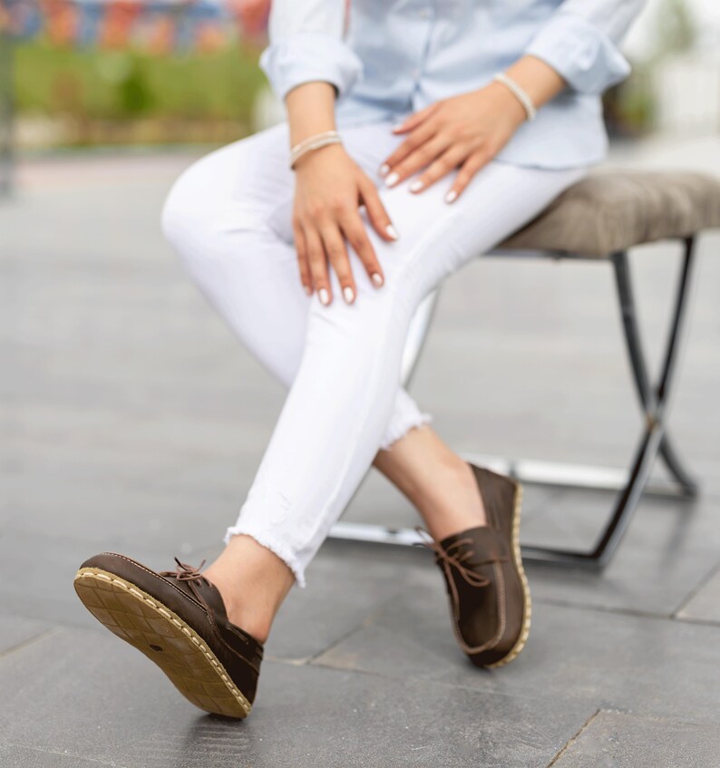 May include: Brown leather boat shoes with light brown stitching and laces. The soles are a light tan color. The shoes are worn with white pants and a light blue shirt.