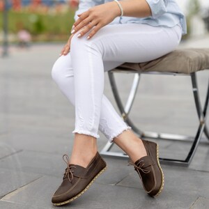 May include: Brown leather boat shoes with light brown laces and stitching. The shoes have a rounded toe and a light-colored sole. The shoes are worn with white pants.