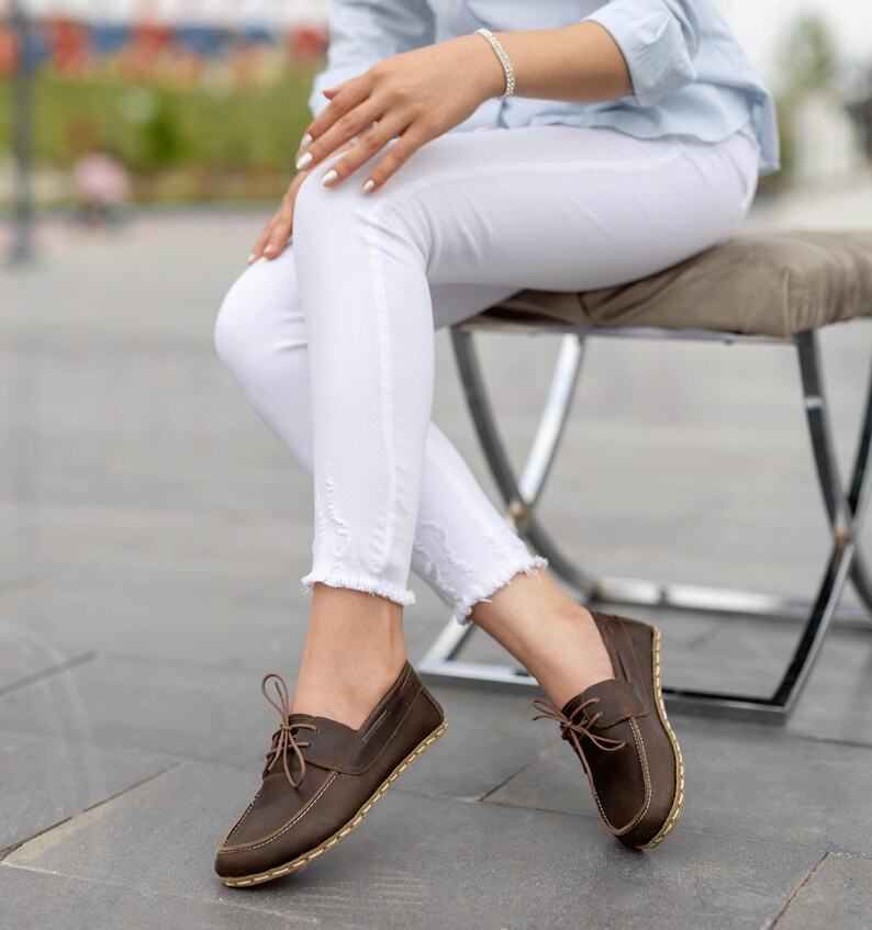 May include: Brown leather boat shoes with light brown laces and stitching. The shoes have a rounded toe and a light-colored sole. The shoes are worn with white pants.