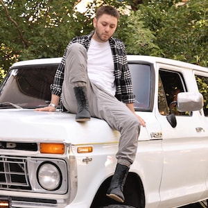 May include: A white Ford F-250 pickup truck with a person wearing a black and white plaid shirt, white t-shirt, gray pants, and black boots. The truck has a round headlight and an orange turn signal.
