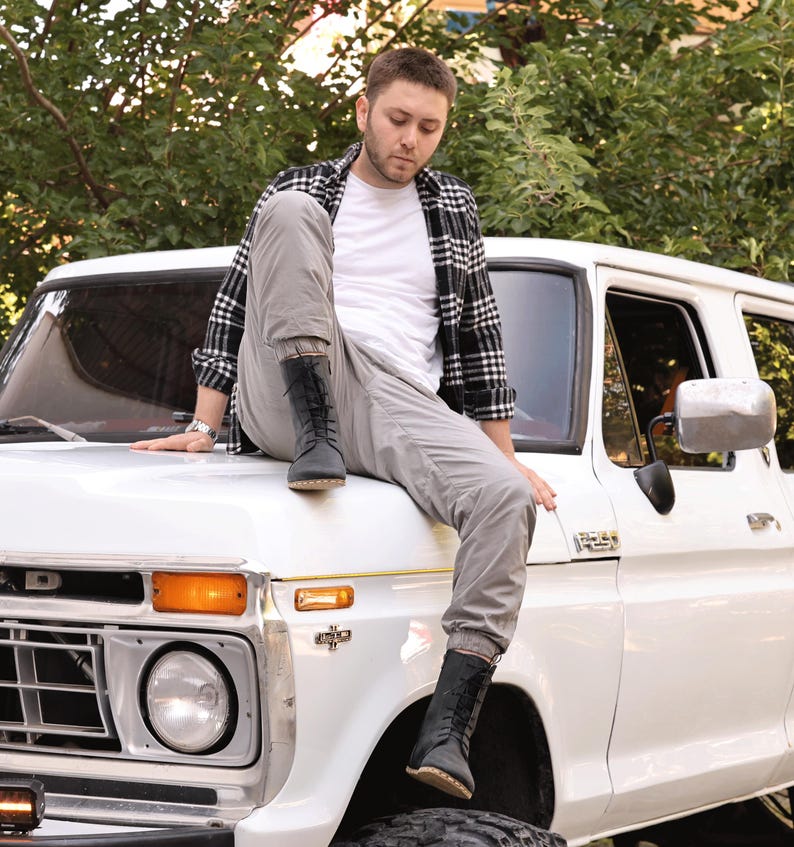 May include: A white Ford F-250 pickup truck with a person wearing a black and white plaid shirt, white t-shirt, gray pants, and black boots. The truck has a round headlight and an orange turn signal.
