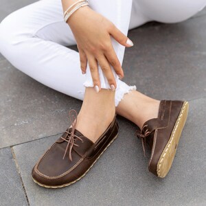 May include: Brown leather boat shoes with light brown laces and stitching. The shoes have a rounded toe and a light-colored sole. The shoes are worn with white jeans.