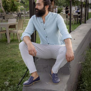 May include: A person wearing a light blue button-down shirt, light gray pants, and blue leather loafers. The person is sitting on a concrete ledge outdoors, with a background of tables and chairs.