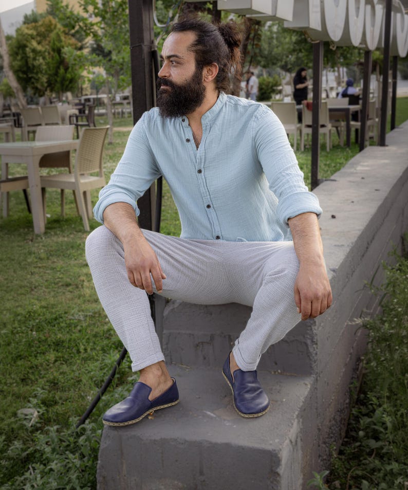 May include: A person wearing a light blue button-down shirt, light gray pants, and blue leather loafers. The person is sitting on a concrete ledge outdoors, with a background of tables and chairs.