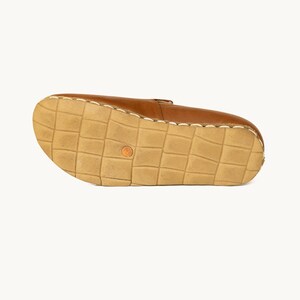 May include: A brown leather shoe, viewed from the sole. The sole is a light tan color with a grid pattern. The shoe's upper is a medium brown, and the stitching is visible along the edges. A small, round, orange-colored detail is on the sole.