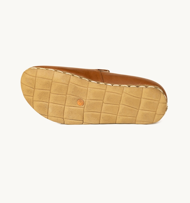 May include: A brown leather shoe, viewed from the sole. The sole is a light tan color with a grid pattern. The shoe's upper is a medium brown, and the stitching is visible along the edges. A small, round, orange-colored detail is on the sole.