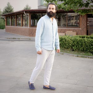 May include: A light blue button-down shirt and light gray pants are worn with navy blue slip-on shoes. The person is standing on a gray paved surface. The shirt has long sleeves and a mandarin collar.