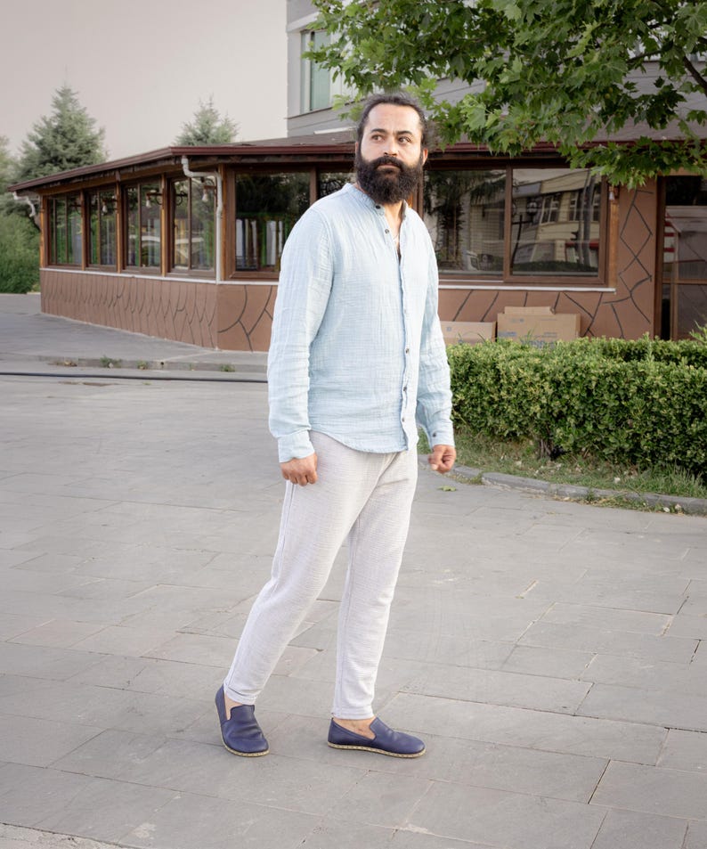 May include: A light blue button-down shirt and light gray pants are worn with navy blue slip-on shoes. The person is standing on a gray paved surface. The shirt has long sleeves and a mandarin collar.