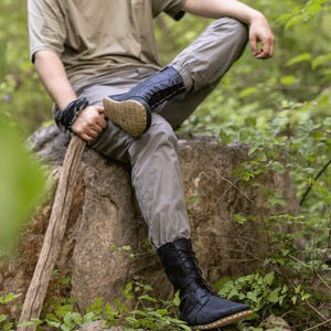 May include: Black leather boots with a lace-up design and a tan sole. The boots are worn by a person sitting on a rock in a forest setting. The boots appear to be made of soft leather.