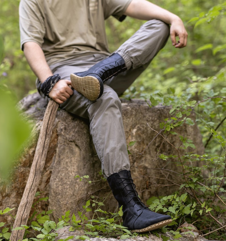 May include: Black leather boots with a lace-up design and a tan sole. The boots are worn by a person sitting on a rock in a forest setting. The boots appear to be made of soft leather.