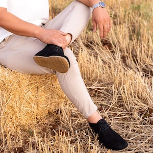 Op de afbeelding: Zwarte suède enkellaarsjes met een bruine zool. De laarzen hebben een vetersluiting en worden gedragen met een beige broek. De persoon zit op een hooibaal. Een horloge is zichtbaar om de pols.