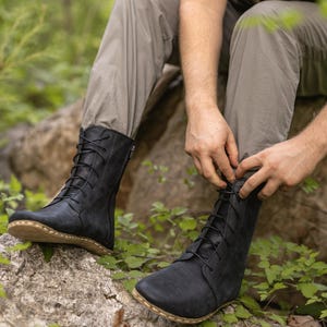 Puede incluir: Botas de cuero negro con cordones y cremallera lateral. Las botas tienen una puntera redondeada y una suela marrón claro. La persona se está atando los cordones de las botas mientras está sentada en una roca. Las botas son parte de una colección de calzado minimalista.
