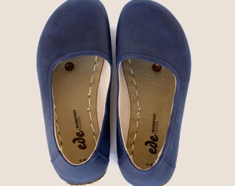 May include: A pair of navy blue slip-on shoes. The shoes have a rounded toe and a stitched sole. The interior is a light tan color with the words "ede BAREFOOT SHOES" printed on the insole. The shoes are size 9W.