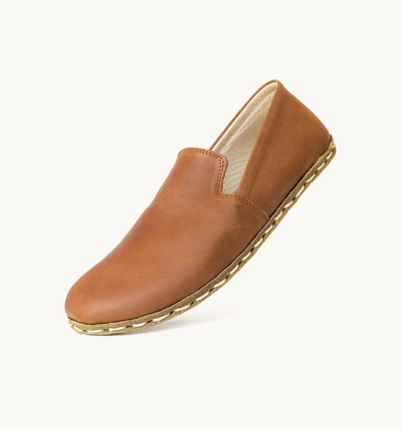 May include: A single brown leather slip-on shoe. The shoe has a rounded toe and a low profile. The sole is stitched with a light-colored thread. The shoe is made of a smooth, polished leather.