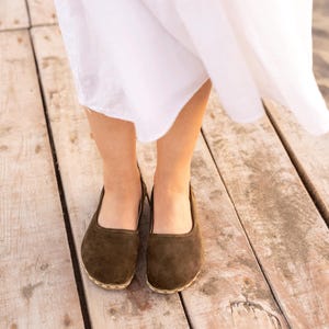 May include: A pair of brown suede flats with a rounded toe and a stitched sole. The shoes are worn on a wooden surface, with a white dress visible above the shoes.
