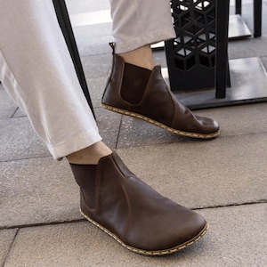 Könnte beinhalten: Braune Leder-Ankle Boots mit elastischen Seiteneinsätzen und einer genähten Sohle. Die Stiefel werden von einer Person getragen, die eine helle Hose trägt. Minimalistisches Design.