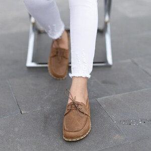 Könnte beinhalten: Braune Lederschuhe mit Schnürsenkeln und weißen Nähten. Die Schuhe werden mit weißen Jeans mit ausgefranstem Saum getragen. Das Bild ist aus einem niedrigen Winkel aufgenommen und zeigt die Schuhe und einen Teil der Beine.