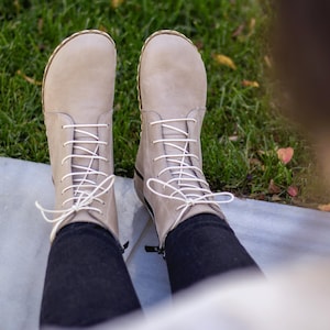 Puede incluir: Botines color beige claro con cordones blancos. Las botas tienen una punta redondeada y una cremallera lateral. Las botas se usan con pantalones negros. El fondo es hierba verde.