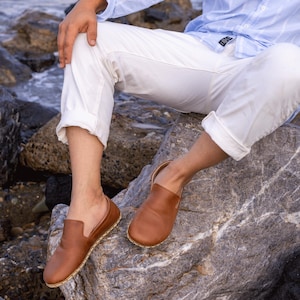 Op de afbeelding: Een paar bruine leren instappers met een ronde neus en een gestikte zool. De schoenen hebben een warme bruine kleur en lijken versleten. De schoenen worden getoond op een persoon die op een rots zit.