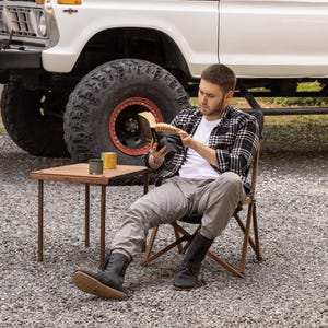 May include: A person seated in a folding chair, reading a book with the title "Livunels". A small wooden table holds two cans. A white truck with large tires is in the background. The person is wearing a plaid shirt, white t-shirt, and gray pants.