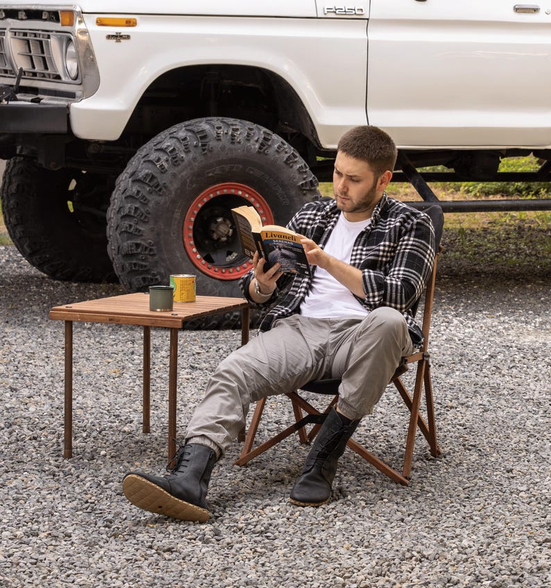 May include: A person seated in a folding chair, reading a book with the title "Livunels". A small wooden table holds two cans. A white truck with large tires is in the background. The person is wearing a plaid shirt, white t-shirt, and gray pants.
