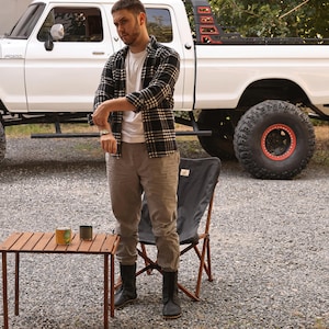 May include: A man stands near a white pickup truck with large tires. He wears a black and white plaid shirt, a white t-shirt, and gray pants. A small wooden table holds two cans. A black folding chair is nearby.