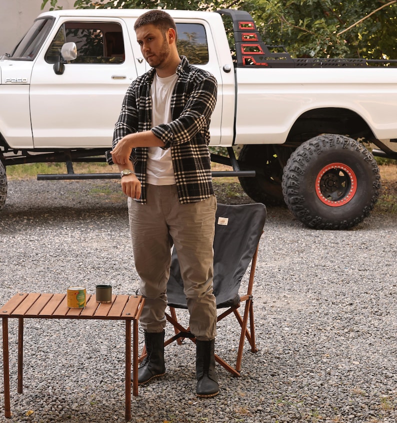 May include: A man stands near a white pickup truck with large tires. He wears a black and white plaid shirt, a white t-shirt, and gray pants. A small wooden table holds two cans. A black folding chair is nearby.