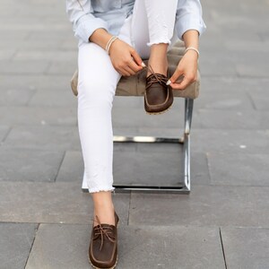 May include: Brown leather boat shoes with light-colored stitching and laces. The shoes have a classic design and are being tied. The image shows the shoes in detail, highlighting their construction and style.