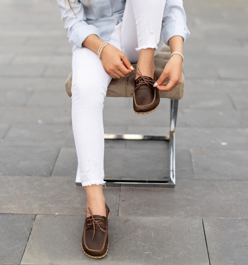 May include: Brown leather boat shoes with light-colored stitching and laces. The shoes have a classic design and are being tied. The image shows the shoes in detail, highlighting their construction and style.