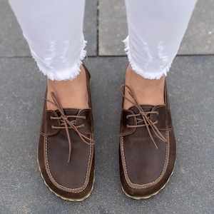 May include: A pair of brown leather boat shoes with light brown laces and stitching. The shoes are worn with white, frayed-hem jeans. The image is taken from a top-down perspective, showcasing the footwear.