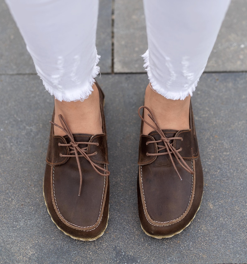 May include: A pair of brown leather boat shoes with light brown laces and stitching. The shoes are worn with white, frayed-hem jeans. The image is taken from a top-down perspective, showcasing the footwear.