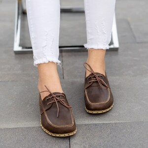 May include: Close-up of brown leather boat shoes with light-colored stitching and laces. The shoes are worn with white, distressed jeans. The shoes have a square toe and a light-colored sole.