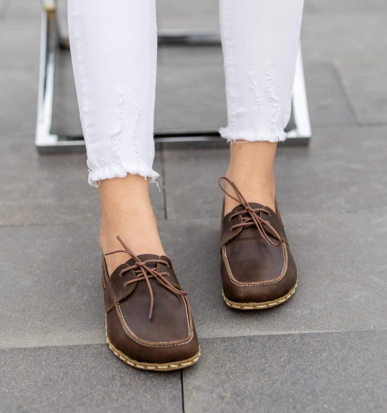May include: Close-up of brown leather boat shoes with light-colored stitching and laces. The shoes are worn with white, distressed jeans. The shoes have a square toe and a light-colored sole.