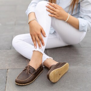 May include: Brown leather boat shoes with light-colored soles and laces. The shoes are worn by a person sitting on a gray surface. The shoes have a classic, casual design.