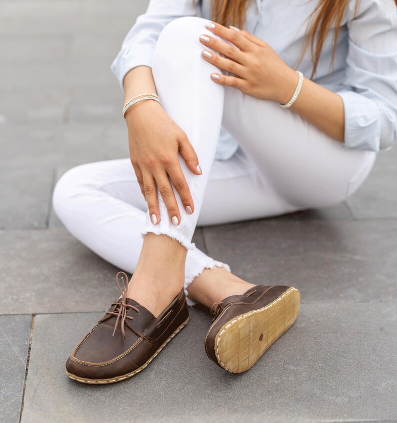 May include: Brown leather boat shoes with light-colored soles and laces. The shoes are worn by a person sitting on a gray surface. The shoes have a classic, casual design.