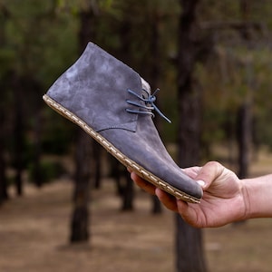 Könnte beinhalten: Ein grauer Wildleder-Knöchelstiefel mit blauen Schnürsenkeln und einer genähten Sohle. Der Stiefel wird in einer Hand vor einem verschwommenen Hintergrund aus Bäumen gehalten. Der Schuh hat ein minimalistisches Design.