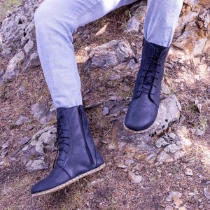 May include: A pair of dark blue leather boots with laces and zippers. The boots have a minimalist design with a natural-colored sole. The boots are worn with light blue jeans and are set against a natural outdoor background.