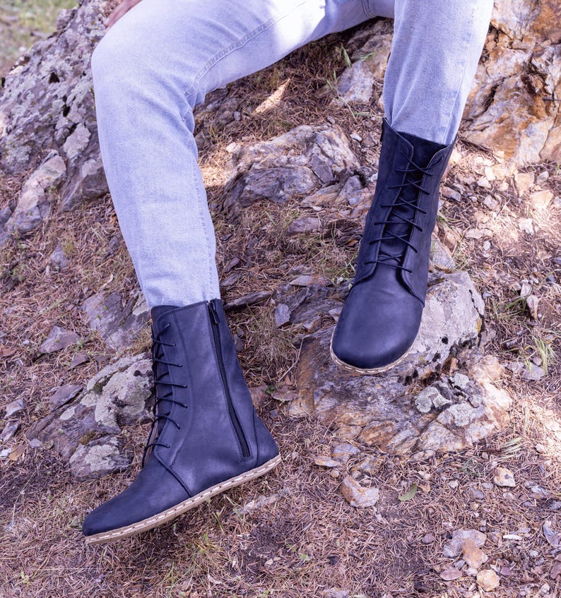 May include: A pair of dark blue leather boots with laces and zippers. The boots have a minimalist design with a natural-colored sole. The boots are worn with light blue jeans and are set against a natural outdoor background.
